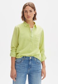 Lime green linen shirt with a collar, button placket, and single chest pocket; worn over a white top, paired with light blue jeans.