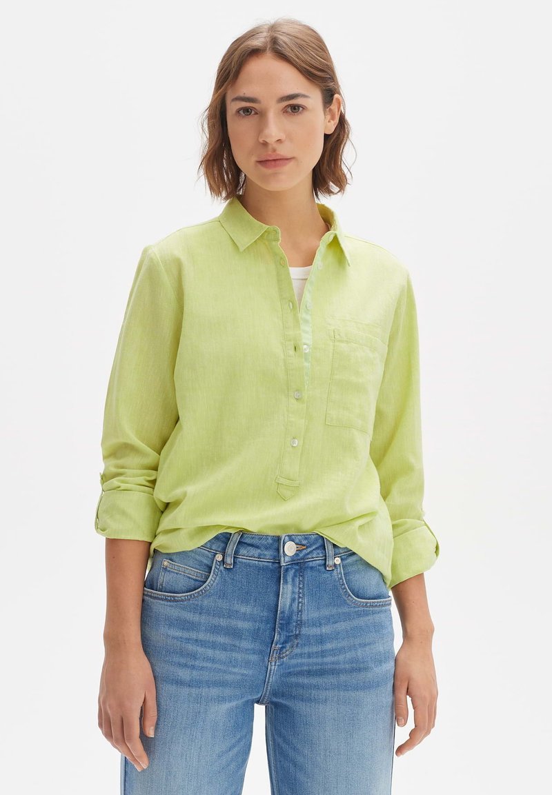 Lime green linen shirt with a collar, button placket, and single chest pocket; worn over a white top, paired with light blue jeans.