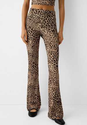 Femme portant un pantalon évasé taille haute à imprimé léopard, assorti à un haut court assorti, et des chaussures plates noires, debout devant un fond blanc.