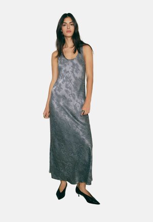 Femeie purtând o rochie gri de satin, fără mâneci, lungime până la gleznă, cu model floral, pantofi negri cu vârf ascuțit și inele pe ambele mâini.