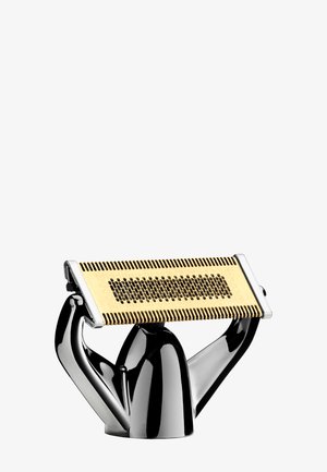 Rasoir électrique or et noir avec une grille, maintenu à la verticale par un support noir brillant et chromé sur un fond blanc.