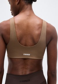 Αθλητικό άτομο που φοράει καφέ αθλητικό σουτιέν με το κείμενο "STRONGER" στην πλάτη, συνδυασμένο με σκούρα καφέ ψηλόμεσα κολάν.