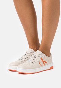 Beiga lädersneakers med perforerade detaljer, orange logotyp på sidan, vit gummisåle och grå snörning. Lågprofil design och vadderad krage.