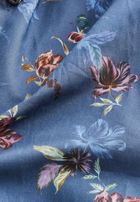 Tissu bleu avec un motif floral comprenant de grandes fleurs et des feuilles dans des tons de rose, violet, jaune et vert, avec bouton et coutures visibles.