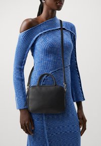 Femme portant une robe côtelée bleue à une épaule avec des détails de boutons et un sac bandoulière en cuir noir à petites poignées.