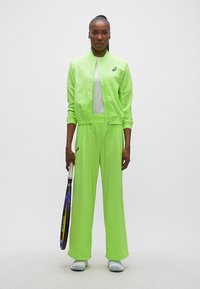 Γυναίκα με φωτεινό πράσινο αθλητικό σετ, κρατώντας ρακέτα τένις, στέκεται μπροστά σε λευκό φόντο.