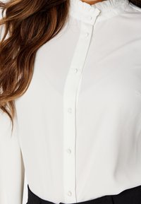 Biała bluzka z długim rękawem, z wysokim falbaniastym kołnierzem i zapinaniem na guziki z przodu. Gładka faktura materiału, dopasowany krój, połączona z czarnym pasem.