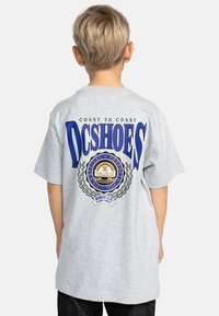 Harmaa puuvillapaita, jossa on suuri sininen DC Shoes -logo takana, jossa on pyöreä emblem ja laakerikuvio. Lyhyet hihat, rento istuvuus.