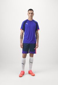 Lila Sporttrikot mit pinken Akzenten, kombiniert mit dunkelblauen Shorts mit einem lila Netzbesatz. Helle orangefarbene Fußballschuhe und weiße Socken.