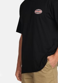 Sort kortærmet t-shirt med et cirkulært logo trykket i rød og hvid på venstre bryst. 100% bomuldsmateriale, afslappet pasform, diskret buet kant.