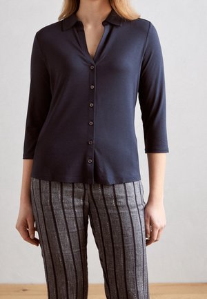Woman wearing a navy blue buttoned top with three-quarter sleeves and gray pants with vertical black stripes.