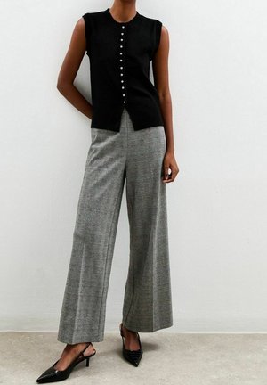 Femme portant un haut noir sans manches à boutons, un pantalon large à taille haute à motif gris à carreaux, et des escarpins noirs à bouts pointus, debout contre un mur blanc.