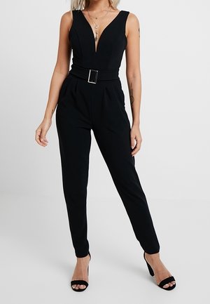 Vrouw in een mouwloze zwarte jumpsuit met een diepe V-hals en een ceintuur in de taille, gecombineerd met zwarte hoge hakken.