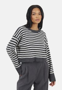 Vrouw in een grijs-wit gestreepte cropped trui met lange mouwen en hoge taille grijze plooienbroek, staand met één hand in de zak.