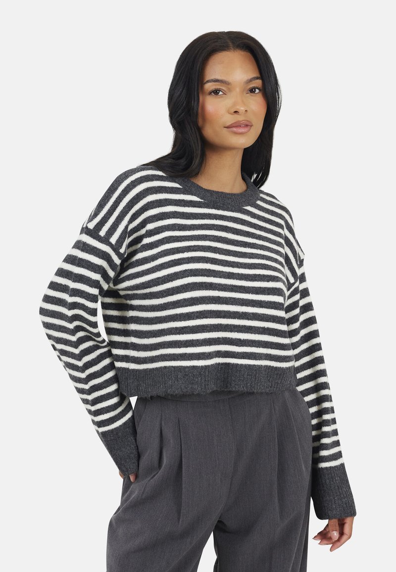 Vrouw in een grijs-wit gestreepte cropped trui met lange mouwen en hoge taille grijze plooienbroek, staand met één hand in de zak.