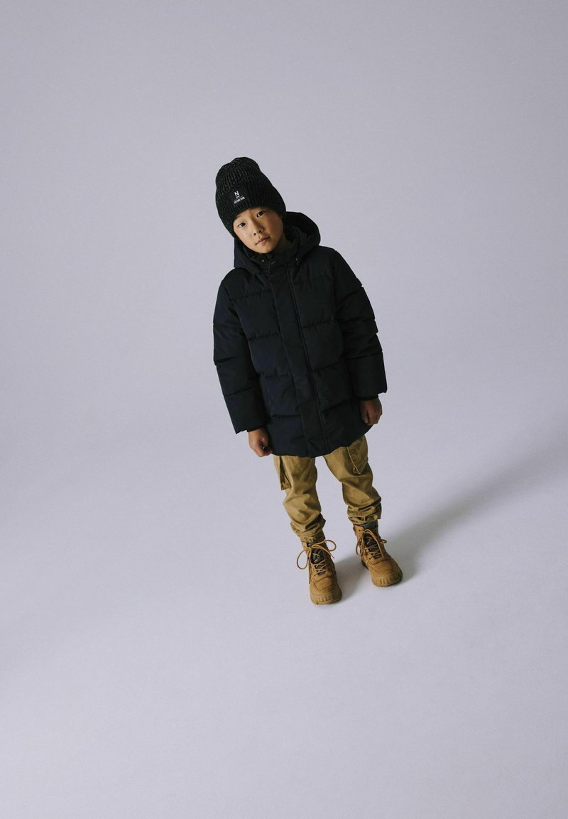 Puffermantel in Schwarz, beigefarbene Cargo-Hosen, braune Stiefel und eine schwarze Strickmütze. Das Kind steht mit neutralem Ausdruck vor einem hellgrauen Hintergrund.