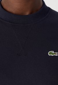 Gros plan d'une personne portant un sweat-shirt bleu marine texturé avec un col côtelé et un petit logo crocodile vert sur la poitrine.