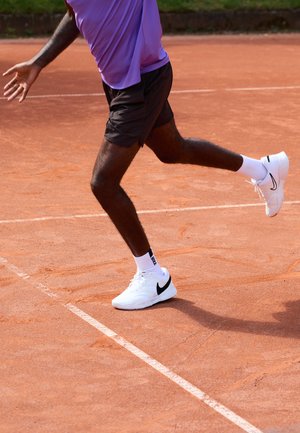 Valkoiset Nike-tennareita mustalla swoosh-logolla, yhdistettynä valkoisiin urheilusukkiin ja mustiin shortseihin, savitenniskentällä.