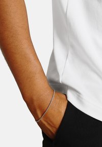 Bracciale in argento con design torcido, indossato su un polso su uno sfondo di camicia bianca e pantaloni neri. Texture liscia e finitura lucida.