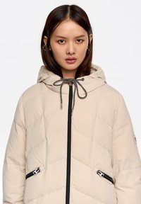 Chaqueta acolchada beige con capucha, con cremallera frontal y bolsillos laterales con cremallera. Cordón ajustable en la capucha.