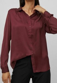 Burgundy silk blouse with long sleeves, button-down front, and a relaxed fit. Smooth texture and slight sheen, paired with black trousers.