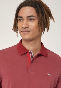 Polo di colore marrone con colletto testurizzato caratterizzato da strisce rosse e blu, chiusura con due bottoni e un piccolo logo ricamato sul petto.
