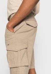 Beige Cargo-Shorts mit seitlichen Taschen und Klappdetails, aus leichtem Material gefertigt. Die Nähte sind entlang der Säume sichtbar, was die Haltbarkeit erhöht.