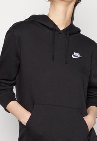 Svart hoodie i mjukt tyg, med en framficka och en liten vit Nike-logotyp på vänster bröst. Justerbar dragsko i huvan.