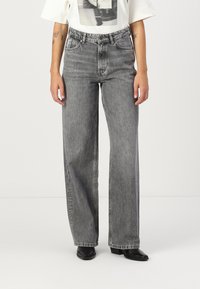 Jeans gri în talie înaltă, cu picioare largi, confecționați din denim, având un finisaj uzat și un design clasic cu cinci buzunare, asortați cu pantofi negri.