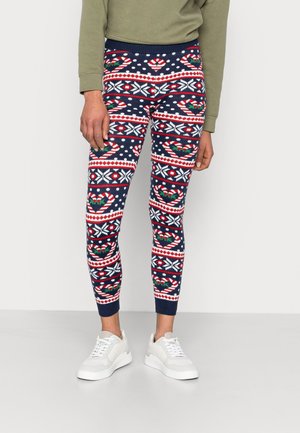 Personne portant des leggings bleu marine avec des motifs de Noël rouges, blancs et verts, des baskets blanches et un haut à manches longues vert olive.