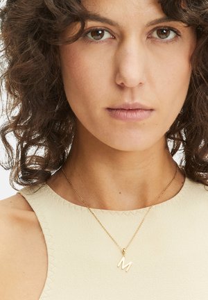 Gouden ketting met een kleine "m" hanger, gedragen op een blootgestelde hals. De ketting heeft een dunne schakel en de omliggende stof is lichtbeige.