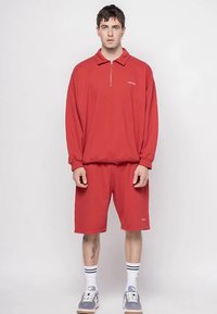 Άνδρας στέκεται με το πρόσωπο προς τα εμπρός, φορώντας κόκκινη φούτερ ζακέτα με φερμουάρ και ταιριαστά κόκκινα σορτς, λευκές κάλτσες και γκρι αθλητικά παπούτσια.