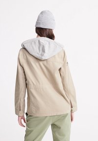 Osoba odwrócona plecami, ubrana w beżową kurtkę z szarym kapturem, szary beanie oraz jasne zielone spodnie, na tle białego tła.