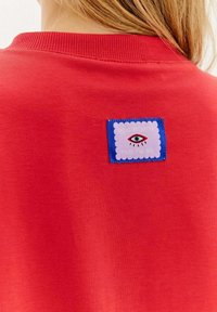 Czerwona bluza z okrągłym kołnierzem, z prostokątną naszywką z niebieską ramką, falistymi krawędziami oraz grafiką oka na plecach.