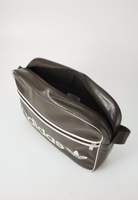 Bruine leren tas met een ritssluiting, witte strepen en zichtbaar Adidas-logo. Binnenkant is van zwart stof met één compartiment.