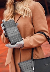 Black patterned wallet featuring a floral design, held in gray-gloved hands, next to a black handbag. Warm brown coat in the background.