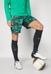 Grünes Langarmshirt, gemusterte grün-schwarze Shorts, schwarze Socken und weiße Fußballschuhe auf einem Fußball mit goldenen Akzenten.