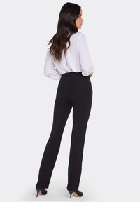 Svarta byxor med hög midja och skräddarsydd passform, med bakfickor, kombinerade med en vit blus, modellen visas bakifrån.