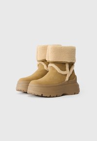 Botas de camurça castanhas com um punho dobrável de pele de carneiro bege, espessa e texturizada. Apresenta uma sola de plataforma robusta com detalhes de tração.