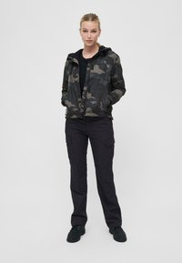 Camo jacket in dark green and black, with a zip front, hood, and pockets, paired with black cargo pants and black shoes.