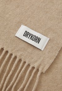 Beige uldsjal med frynser; har et hvidt mærke mærket "DRYKORN" i sort tekst. Blød tekstur, enkelt design.