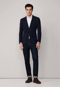 Navyblazer, getailleerde pasvorm, enkele knoop aan de voorkant, gecombineerd met een wit overhemd en donkere spijkerbroek, bruine schoenen en opgerolde mouwen.