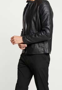 Giacca di pelle nera con colletto alto, chiusura con zip e cuciture verticali. Presenta due tasche laterali e una consistenza liscia.