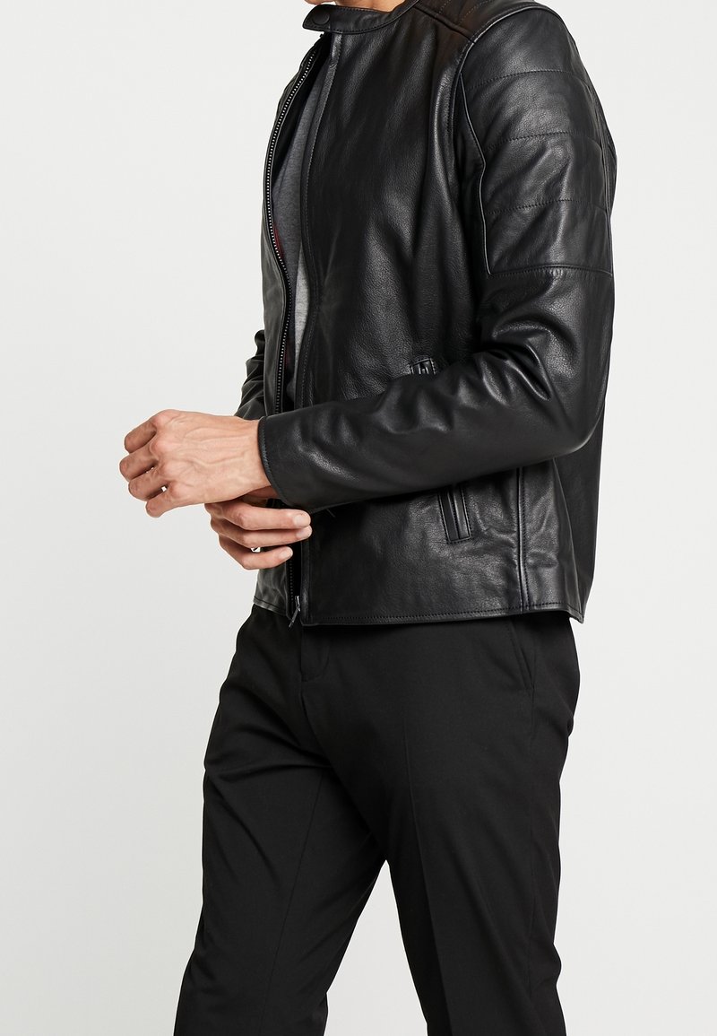 Giacca di pelle nera con colletto alto, chiusura con zip e cuciture verticali. Presenta due tasche laterali e una consistenza liscia.