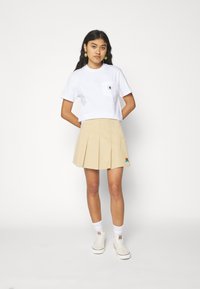 Hvit kortermet T-skjorte med brystlomme, sammen med en beige plisserte skjørt med fargerik broderidetalj. HVite sokker og sneakers.