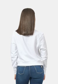 Felpa bianca a maniche lunghe, collo tondo e polsini a costine, indossata con jeans aderenti blu scuro. Tessuto liscio, senza motivi o dettagli visibili.