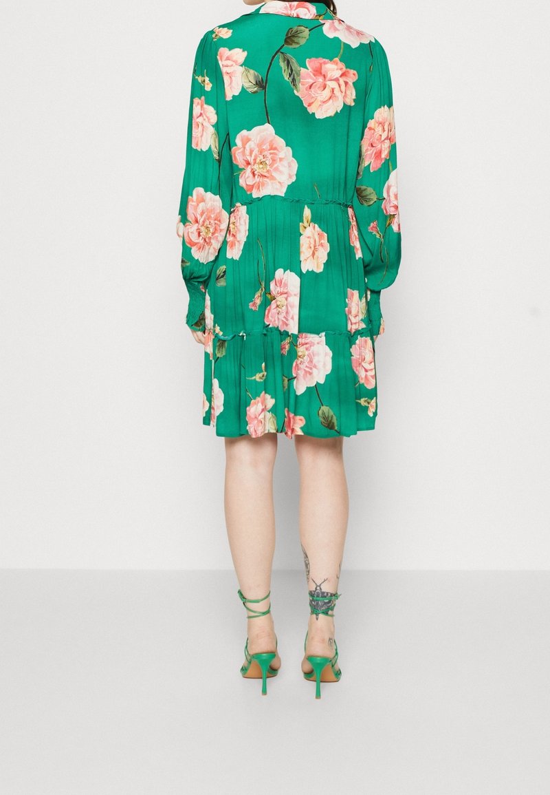 Femme portant une robe verte à fleurs à manches longues et des sandales vertes à talons assorties, debout avec les jambes visibles de dos.