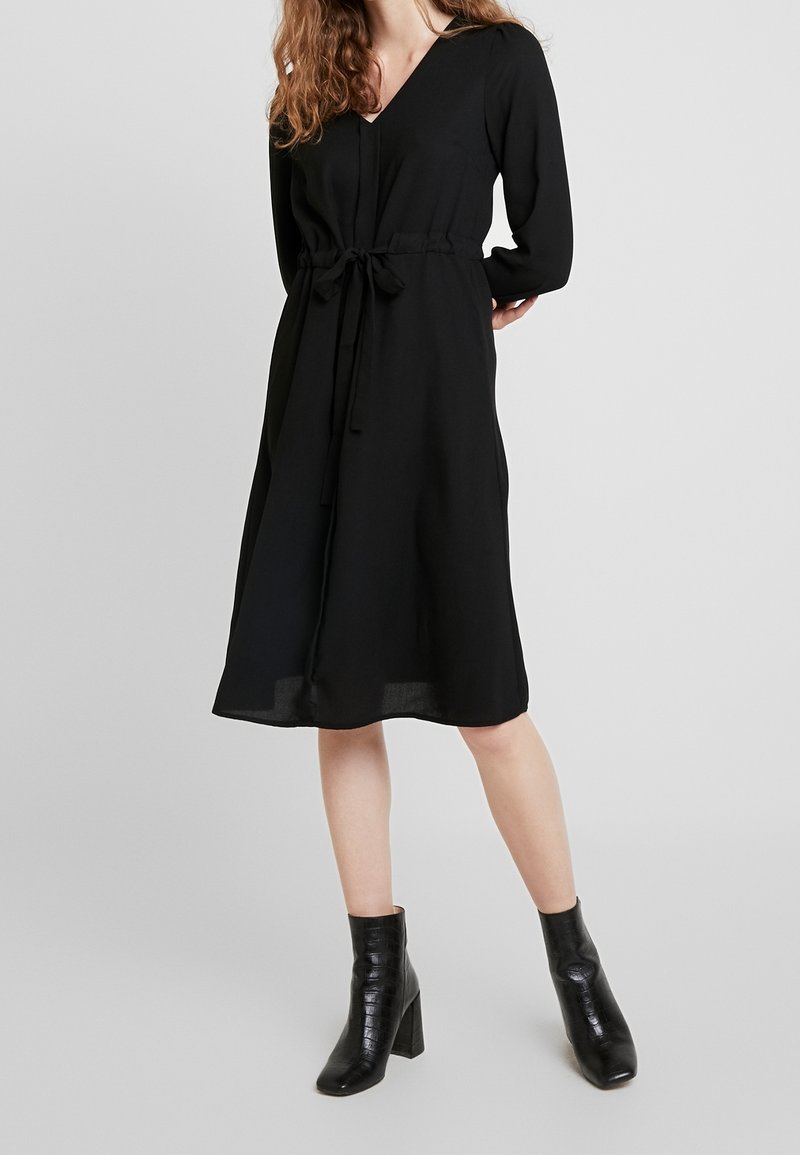 Femme portant une robe midi noire à manches longues avec une ceinture à la taille, associée à des bottines noires à talons, debout devant un fond uni.