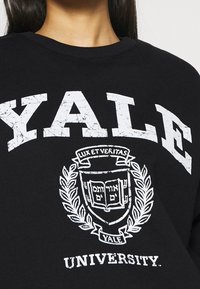 Felpa nera con la scritta "YALE" in grandi lettere bianche usurate, con un emblema di Yale e "UNIVERSITY" sottostante in bianco. Tessuto morbido.