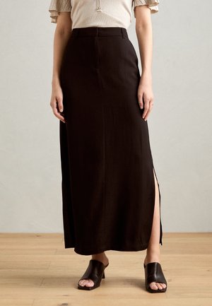 Jupe noire longue jusqu'au sol avec une fente latérale, taille cintrée et tissu lisse. Associée à des mules noires à talons ouverts sur un sol en bois.